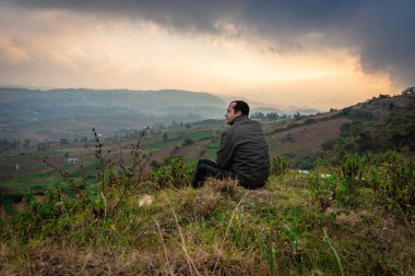Dağın tepesinde oturan genç adam sisli tepe arkaplanı ve sabah dramatik gökyüzü ile Badda Peak Shillong Meghalaya Hindistan 'da çekilmiştir..
