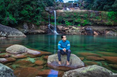 Genç adam, el değmemiş şelale arkaplanlı bir kayanın üzerinde sabahleyin ormanlarda görüntüler çekiliyor..