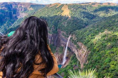 Nohkalikai Şelalesi Cherrapunji Meghalaya Hindistan 'da gündüz vakti yeşil ormanlarla kaplı dağların tepesinden akan şelaleleri seyreden genç bir kız görülüyor..