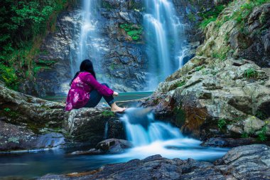 Şelaleyle dağdan düşen ve sabah bulanık su yüzeyi olan kayanın üzerinde oturan kız, Şillong Meghalaya Hindistan 'da çekiliyor..