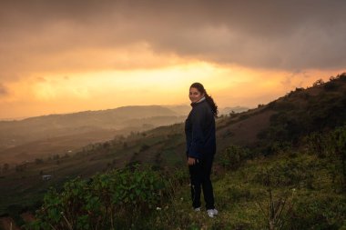 Sisli tepe arkaplanı ve sabah dramatik gökyüzü ile dağın tepesinde duran genç kız Badda Peak Shillong Meghalaya Hindistan 'da çekilmiştir..