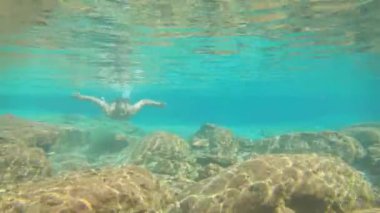 Genç adam maviler içinde yüzüyor. Şelalenin suları altında. Düz açılı pek çok kaya var.