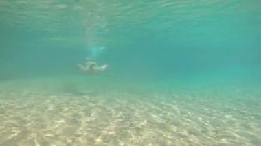 Genç adam maviler içinde yüzüyor. Şelalenin suları altında. Düz açılı pek çok kaya var.