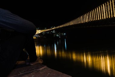 Feribot ışıklarıyla aydınlatılan su yansıtmalı demir asma köprüsünü izleyen adam, Rishikesh uttrakhand Hindistan 'da çekildi..