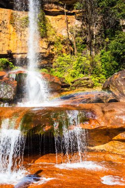 Şelalenin beyaz akıntıları dağın tepesinden düşüyor. Sarı kayalıklarda sabah gökkuşağı var.