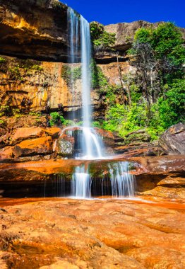 Şelalenin beyaz akıntıları dağın tepesinden düşüyor. Sarı kayalıklarda sabah gökkuşağı var.