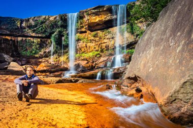 Şelalenin yanında oturan izole genç adam mavi gökyüzüyle dağın tepesinden düşüyor.