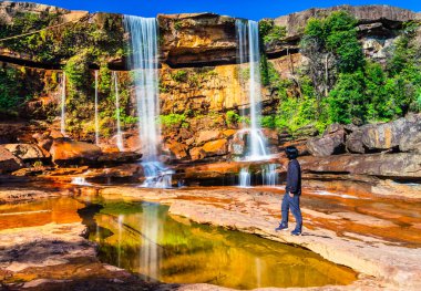 Yansıması ve mavi gökyüzü ile dağdan düşen şelaleyi izleyen izole genç adam.