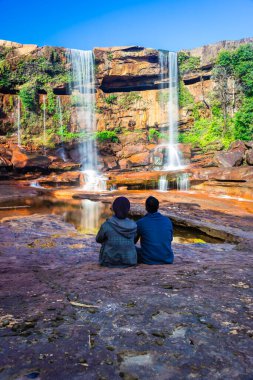 Şelalenin yanında oturan izole genç adam, yansıması ve mavi gökyüzü ile dağın tepesinden düşüyor.