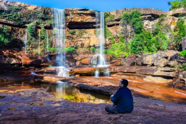 Şelalenin yanında oturan izole genç adam, yansıması ve mavi gökyüzü ile dağın tepesinden düşüyor.