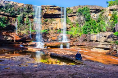 Yalnız genç adam, şelalenin yakınlarında meditasyon yapıyor. Dağların tepesinden düşüyor. Yansıması ve mavi gökyüzü ile.