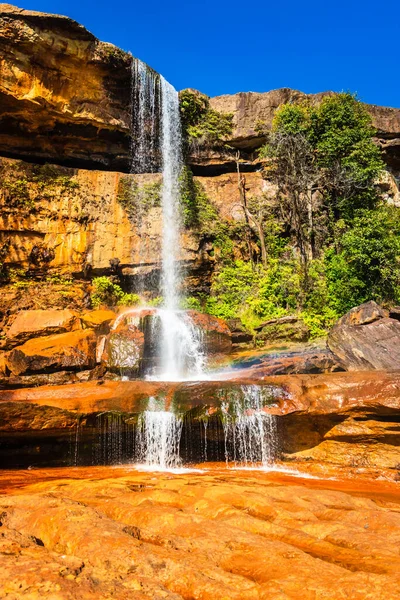Şelalenin beyaz akıntıları dağın tepesinden düşüyor. Sarı kayalıklarda sabah gökkuşağı var.