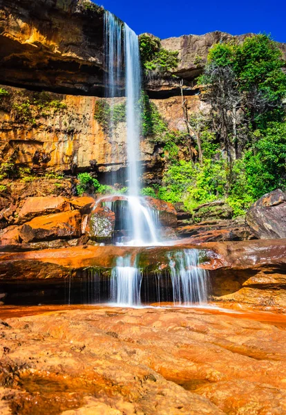 Şelalenin beyaz akıntıları dağın tepesinden düşüyor. Sarı kayalıklarda sabah gökkuşağı var.