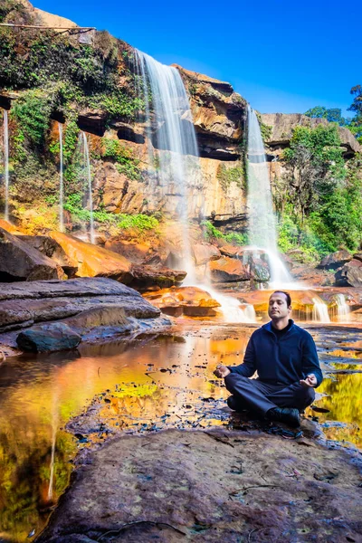 Yalnız genç adam, şelalenin yakınlarında meditasyon yapıyor. Dağların tepesinden düşüyor. Yansıması ve mavi gökyüzü ile.