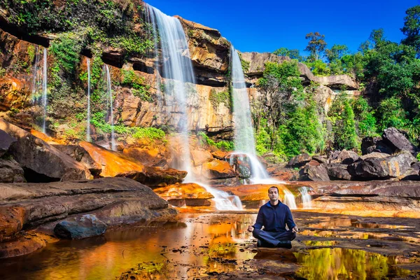 Yalnız genç adam, şelalenin yakınlarında meditasyon yapıyor. Dağların tepesinden düşüyor. Yansıması ve mavi gökyüzü ile.