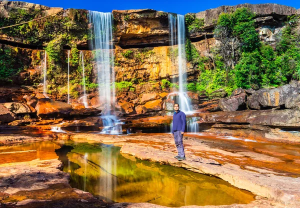Şelalenin yanında oturan izole genç adam, yansıması ve mavi gökyüzü ile dağın tepesinden düşüyor.