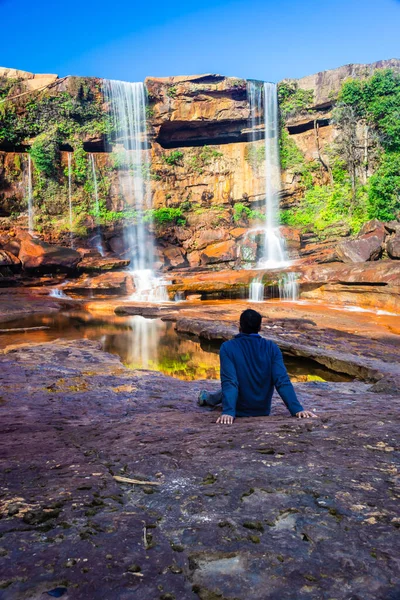 Şelalenin yanında oturan izole genç adam, yansıması ve mavi gökyüzü ile dağın tepesinden düşüyor.