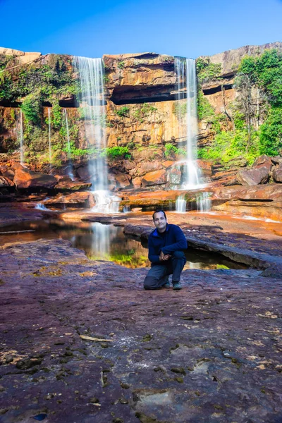 Şelalenin yanında oturan izole genç adam, yansıması ve mavi gökyüzü ile dağın tepesinden düşüyor.