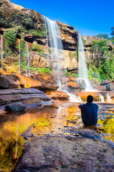 Şelalenin yanında oturan izole genç adam, yansıması ve mavi gökyüzü ile dağın tepesinden düşüyor.