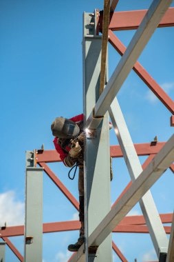 Kaynakçı, mavi bulutsuz gökyüzünün arka planına karşı metal bir yapının üzerinde dikilirken kaynakçılık işi yapar.