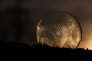 Frozen soap bubble on the snow with ice pattern on cold winter day lighted by the sunset.