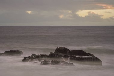 Alacakaranlıkta uzun deniz manzarası ve ön planda ilginç kayalar.
