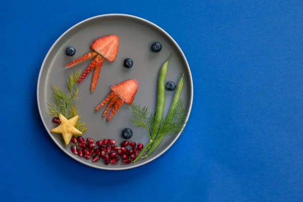 イチゴ ブラックベリー ザクロから海の世界のクラゲ 緑の植物やヒトデ 青い背景を持つプレート上に隔離されたカラフルな食べ物のアイデア — ストック写真