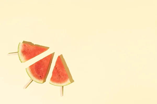 Watermelon pieces with popsicle sticks on a light yellow background. Minimal summer fruit concept. Flat lay.
