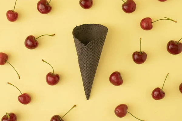 Summer creative pattern with fresh red cherries and black ice cream cone on pastel yellow background. Minimal summer food idea.