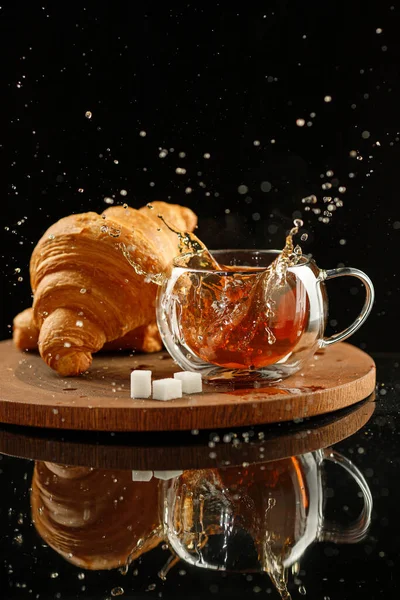 Tea in a glass cup with a splash, croissants and sugar cubes on a wooden holder on black background. — Stock Photo, Image