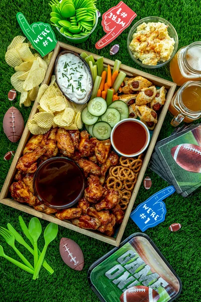 Football tailgate themed party food display with chicken wings, sauces, dips, wieners, vegetables, chips, pretzels, football themed plates, napkins and decorations shot overhead on grass background