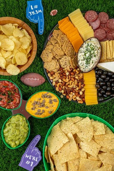 Football tailgate themed party food display with bowls of chips, platter of cheese, crackers, olives, nuts, salami and nuts with spinach articoke dip, salsa, cheese dip and guacamole, portrait orientation