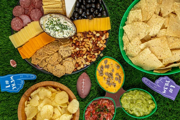 Football tailgate themed party food display with bowls of chips, platter of cheese, crackers, olives, nuts, salami and nuts with spinach articoke dip, salsa, cheese dip and guacamole, horizontal orientation