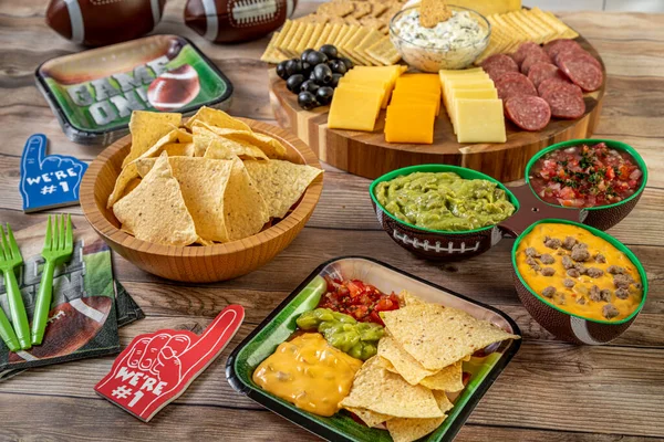 Football party food display with tortilla chips, guacamole, salsa, and queso dips and meat, cheese and cracker tray on wooden table ready for game day celebration