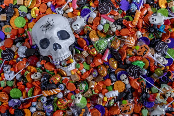 Scary Halloween skull with mouth open and filled with candy and a black spider on forehead, sitting in an assortment of Halloween candy spread out on table top