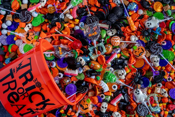 Orange candy bucket with black printed Trick or Treat words on side and assortment of Halloween candy spilling out onto table
