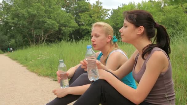 Two female friends drink water, relax and talk after running and doing sport activity in the park — Stock Video