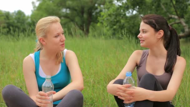 Две подруги пьют воду, отдыхают и разговаривают после занятий спортом в парке — стоковое видео