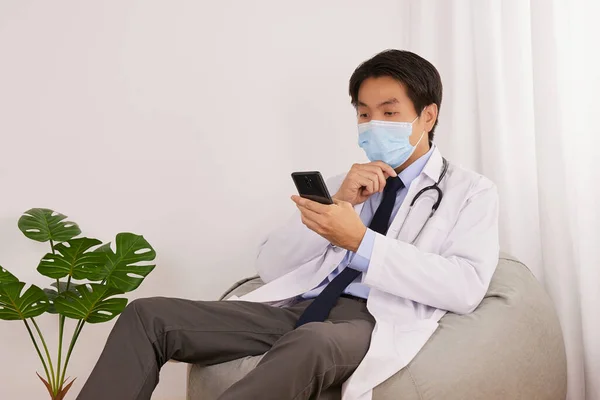 Asian Doctor or Physician Wear Face Mask Serious Thinking and Use Smartphone or Mobile Phone to Check Patient Healthcare in Medical Office or Doctor Room