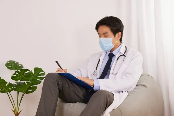 Asian Doctor or Physician Wear Face Mask and Writing Healthcare and Physical Examination Report of Patient at Medical Office or Doctor Room