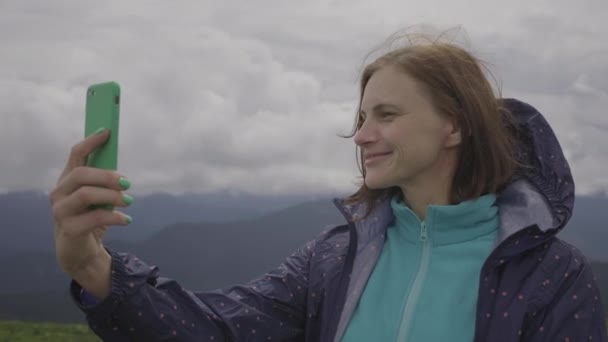 Viajante mujer de 30 años tomando foto selfie en el teléfono celular de pie en las montañas nubladas. Tomar fotos del paisaje. — Vídeo de stock