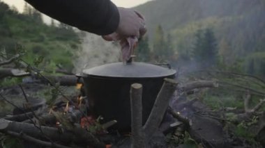 Dağ kampında erkek el, şenlik ateşinin kapağını açıyor. Yanan buharla kaynar su. Dışarıda turist yemeği var.. 