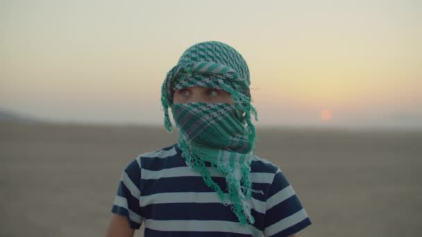 Portret van een jongen met een geruite keffiyeh in de woestijn bij zonsopgang. Woestijnzonsopgang met toeristisch kind. — Stockvideo