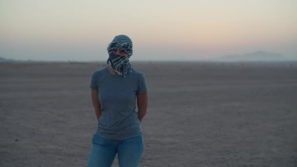 Kobieta ubrana w keffiyeh stojąca na pustyni i czekająca na wschód słońca patrząca w kamerę. Biała kobieta turystka stojąca na pustyni. — Wideo stockowe