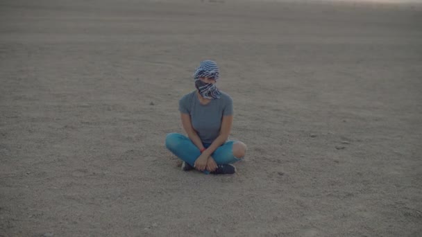 Een vrouw in Checked Keffiyeh zit in de woestijn naar de camera te kijken. Blanke vrouwelijke toerist zit op woestijn zand. — Stockvideo
