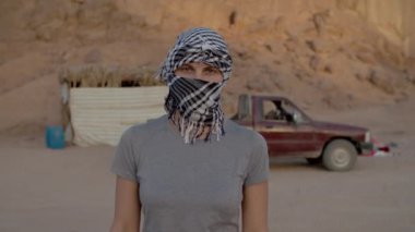 Çölde dikilip kameraya bakan siyah beyaz keffiyeh 'li kadın portresi. Beyaz kadın turist başlığını çıkarıyor ve kameraya gülümsüyor..
