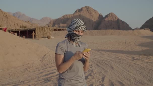 체크인된케피예 여성 관광객 이 핸드폰을 사용하며 사막에 서 있습니다. 카프카스 출신 여성, 사막의 카메라 앞에서 머리쓰개를 벗고 웃으며. — 비디오