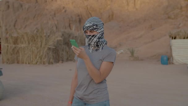 Vrouwelijke toerist in geruite keffiyeh staat in de woestijn met behulp van mobiele telefoon. — Stockvideo