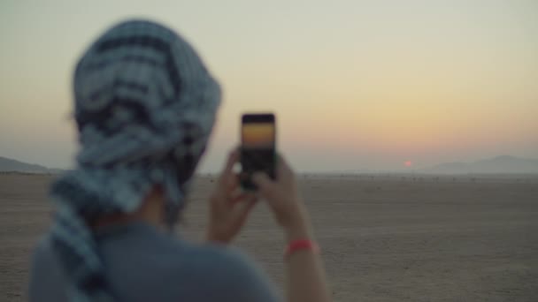 Женщина в клетчатой кеффие фотографирует восход солнца по мобильному телефону, стоящему в пустыне. Кавказские туристки наслаждаются пустынными приключениями. — стоковое видео