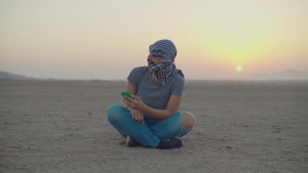 Vrouw in geruite keffiyeh zittend op woestijn zand met behulp van mobiele telefoon. Blanke vrouwelijke toerist genieten van woestijn avontuur tijdens zonsopgang. — Stockvideo